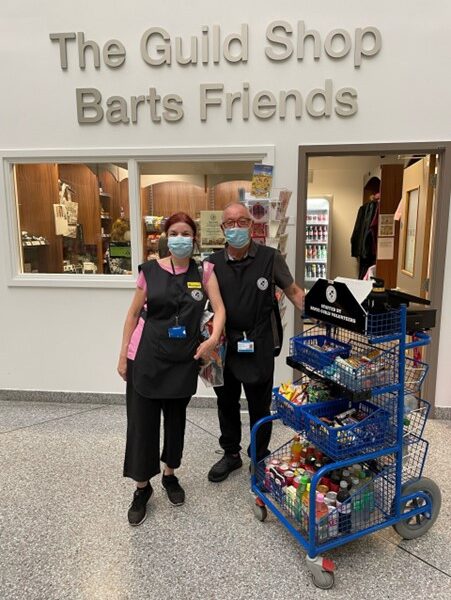 Image of two people standing outside the entrance to the Friends shop inside the hospital, with a laden ward trolley beside them