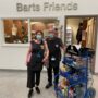 Image of two people standing outside the entrance to the Friends shop inside the hospital, with a laden ward trolley beside them